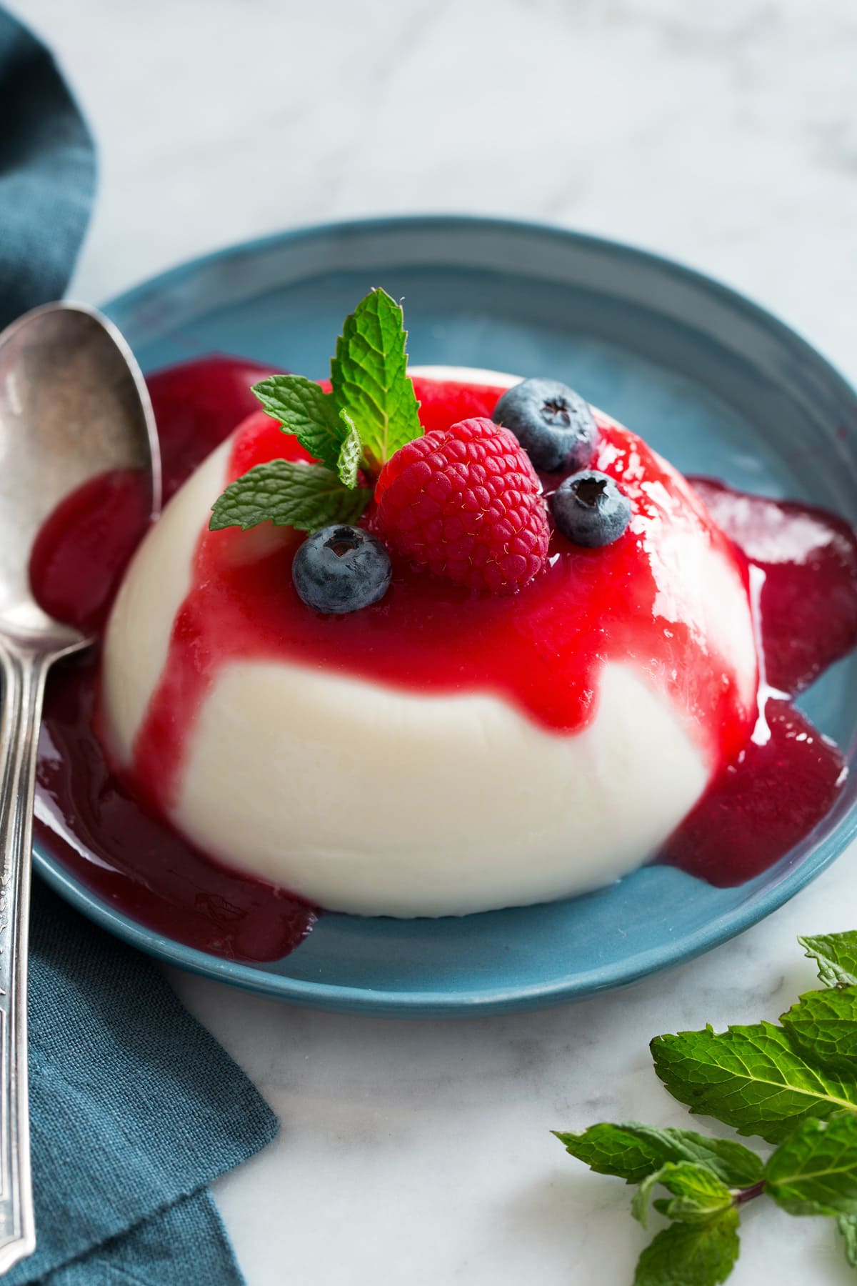 Panna Cotta Photo: Single serving of a disk of Panna Cotta shown on a blue plate covered with raspberry sauce, fresh blueberries and mint.