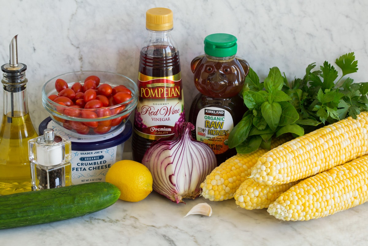Corn Salad Photo: Ingredients used to make corn salad shown in image. Includes corn on the cob, cucumber, red onion, parsley, basil, tomatoes, feta, red wine vinegar, lemon, honey, olive oil, garlic, salt and pepper.
