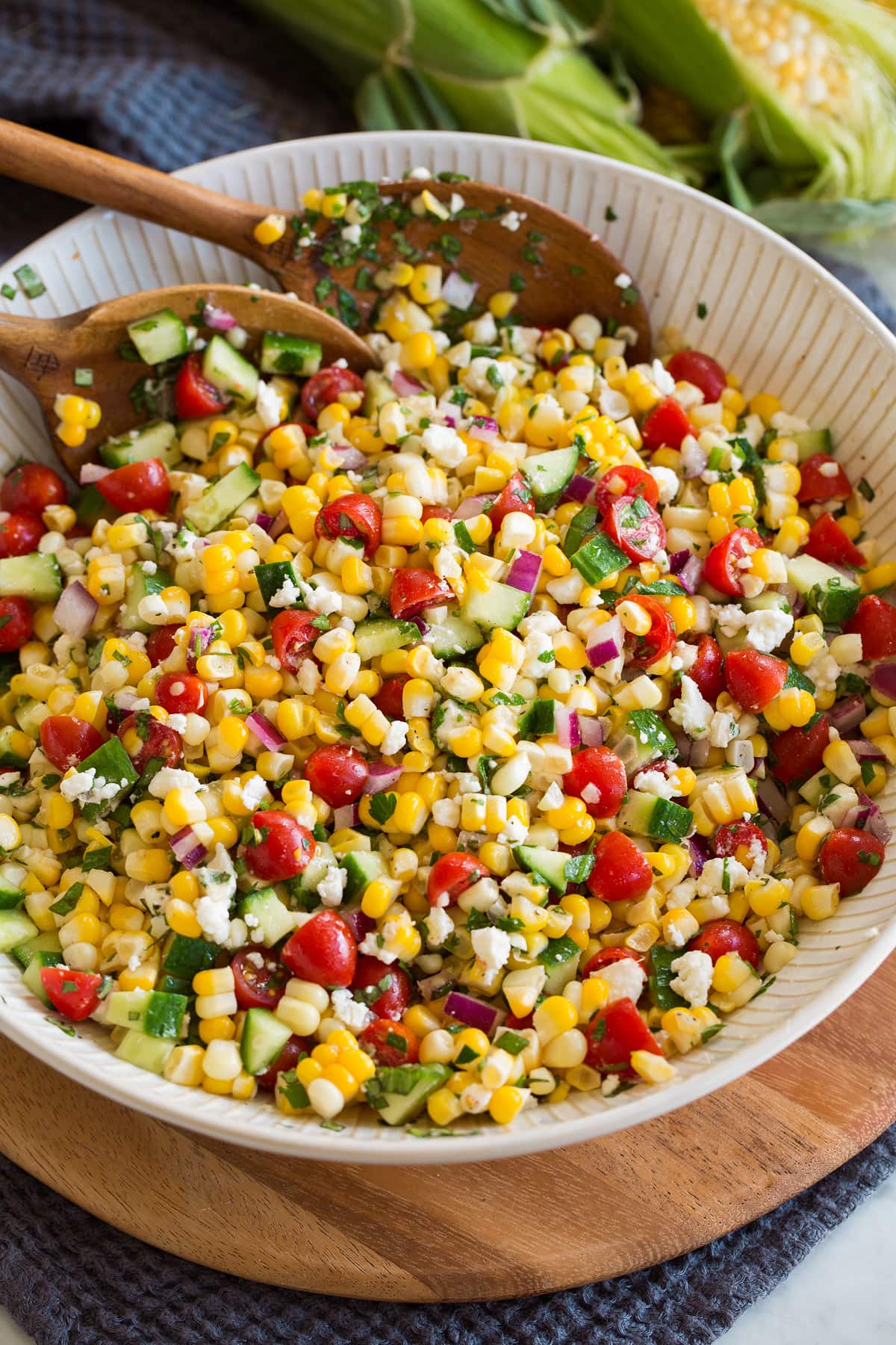 Corn Salad Photo: Corn salad shown in a white serving bowl over a wooden platter and blue cloth. Salad includes fresh corn, tomatoes, red onion, cucumber, feta, herbs and dressing.