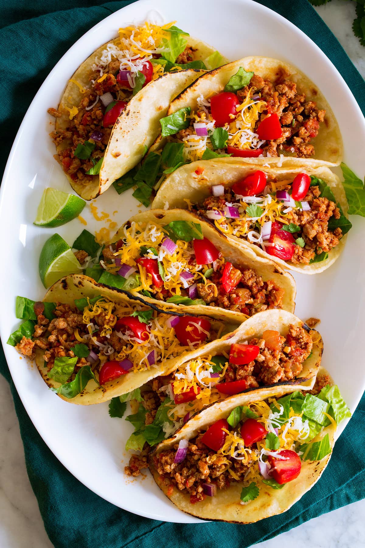 Ground Turkey Tacos Photo: 7 ground turkey tacos in a row shown on a white oval platter from above. Tacos are filled with meat, tomatoes, lettuce, cheese and red onion.