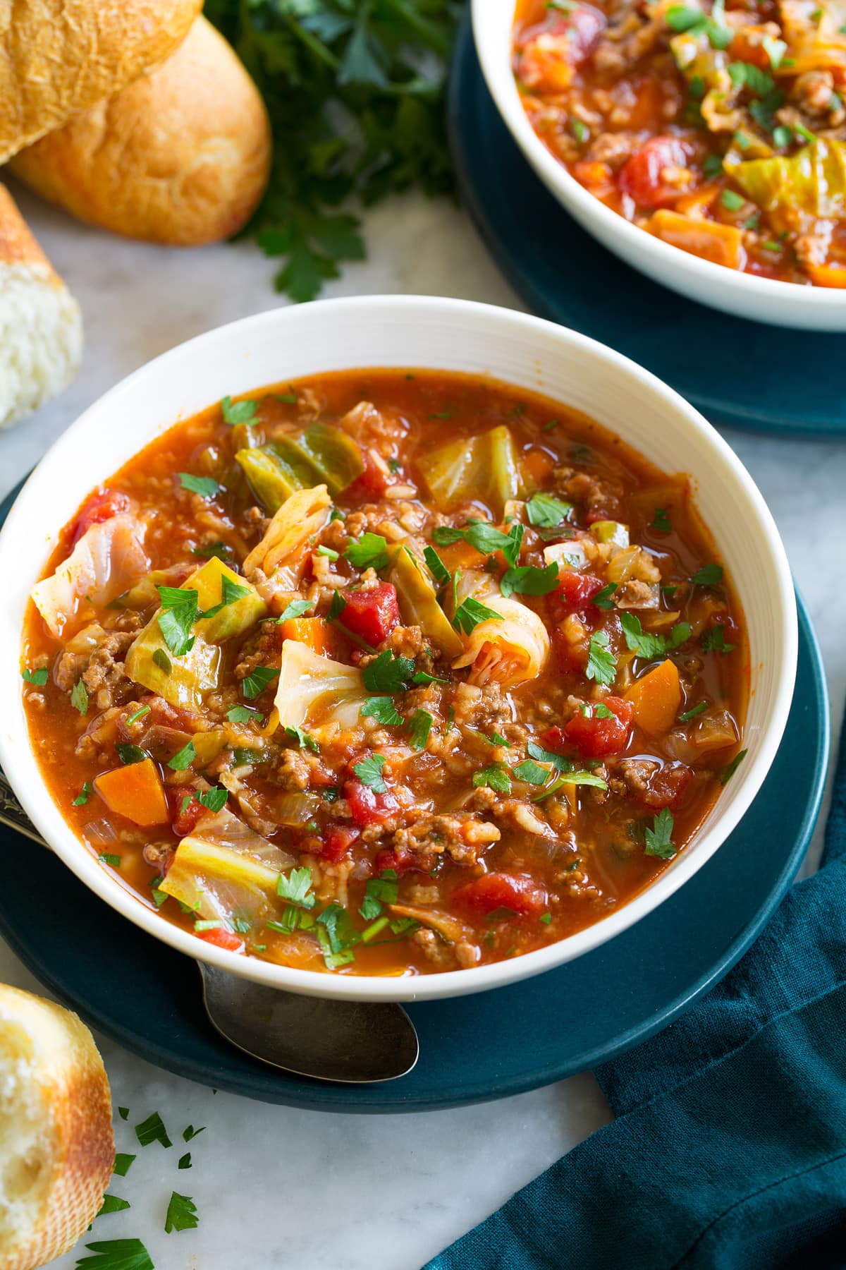Cabbage Roll Soup Single serving of cabbage roll soup in a white bowl set over a blue plate with fresh bread shown as a serving suggestion on the side.