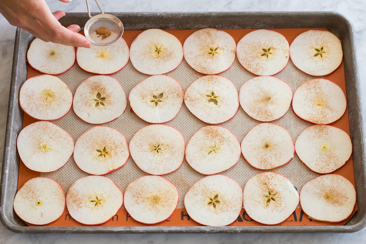 Baked Apple Chips Thin apple slices on a silicone baking mat lined baking sheet being dusted with cinnamon.