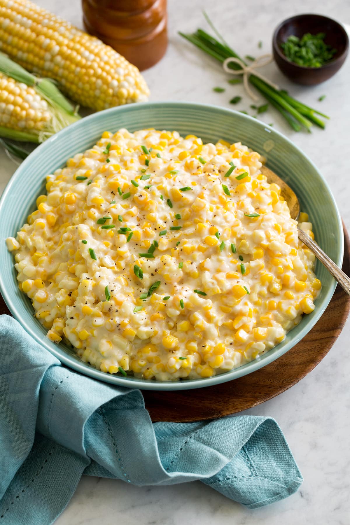 Creamed Corn Creamed corn in a turquoise serving bowl garnished with chives and pepper. Bowl is resting on a wood plate with a blue cloth to the side.