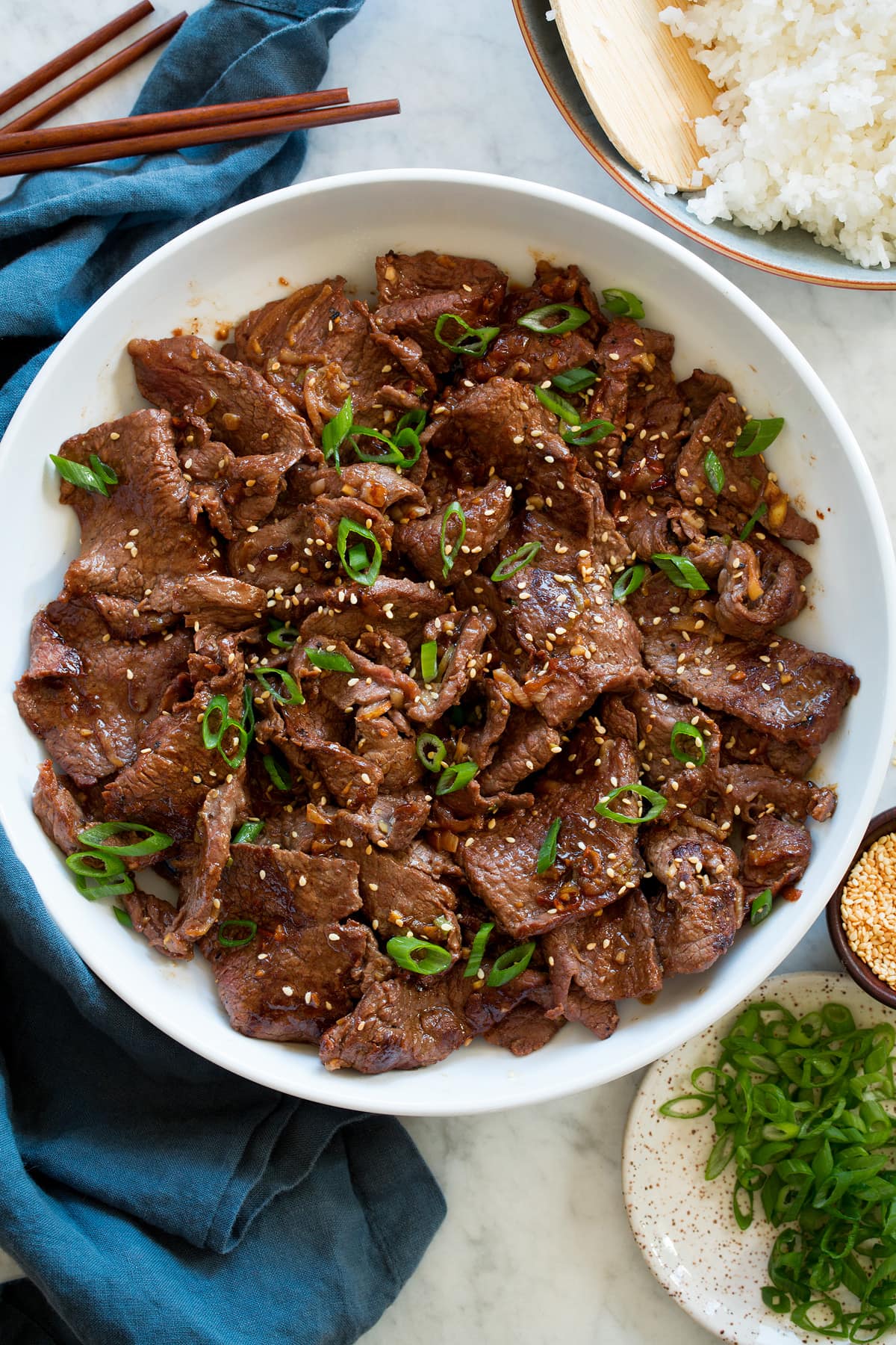Bulgogi Bulgogi shown overhead in a white serving bowl with wooden chopsticks, rice, green onions and a blue cloth to the side.