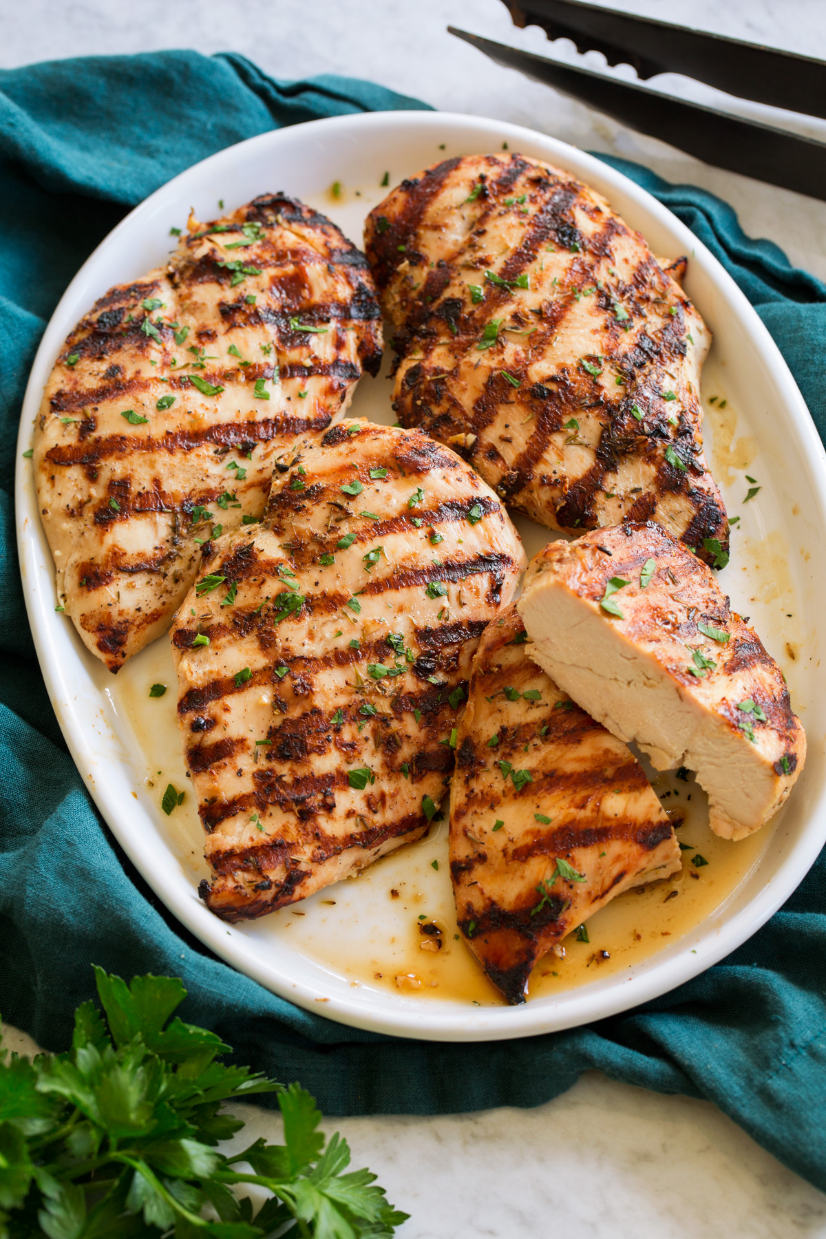 Grilled Chicken Breasts Grilled Chicken Breasts on a white platter on a teal colored cloth shown from a side angle with parsley and grilling tongs to the side.