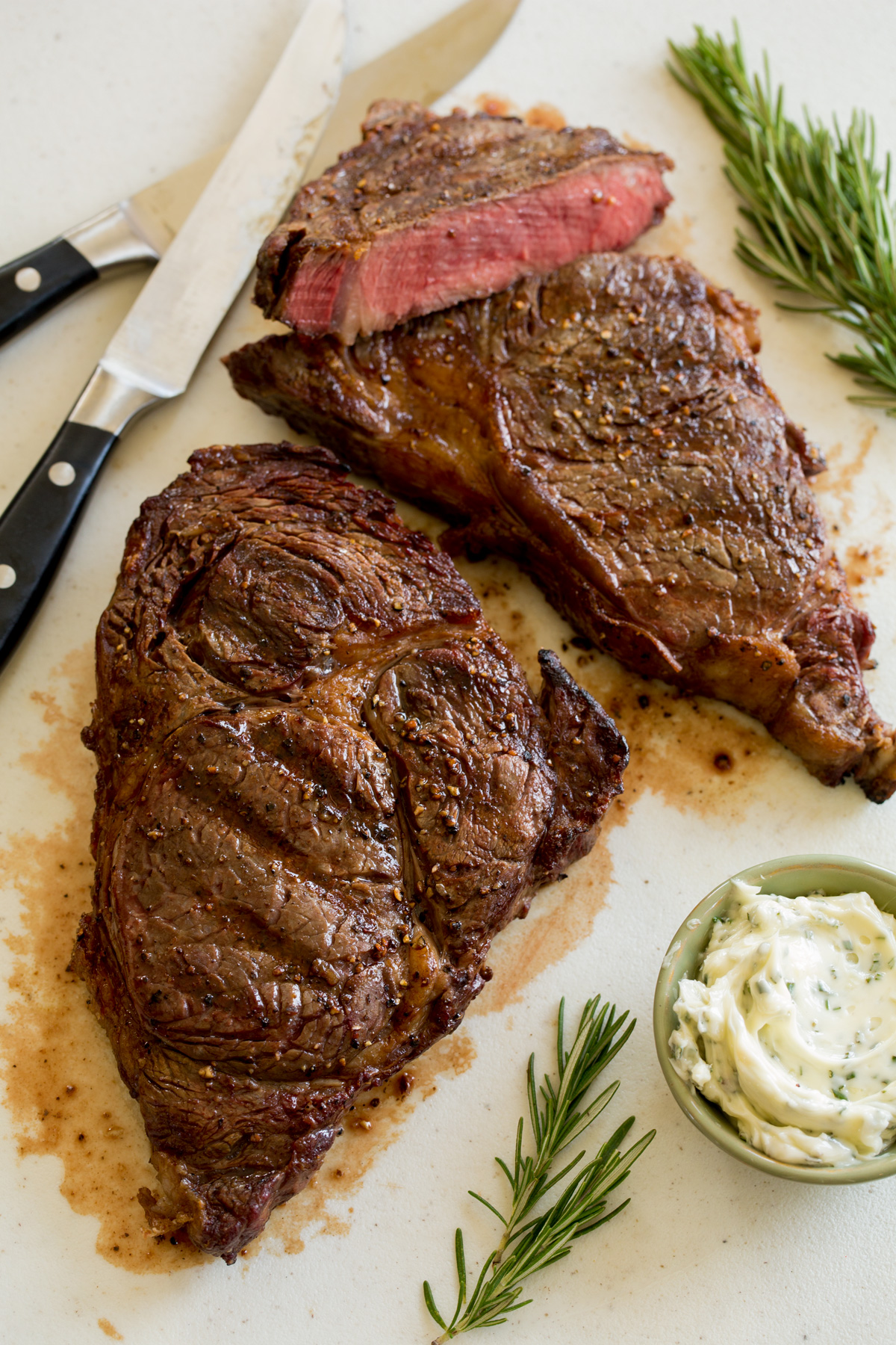 Grilled Steak Two grilled steaks on a cutting board with knives and rosemary to the side.