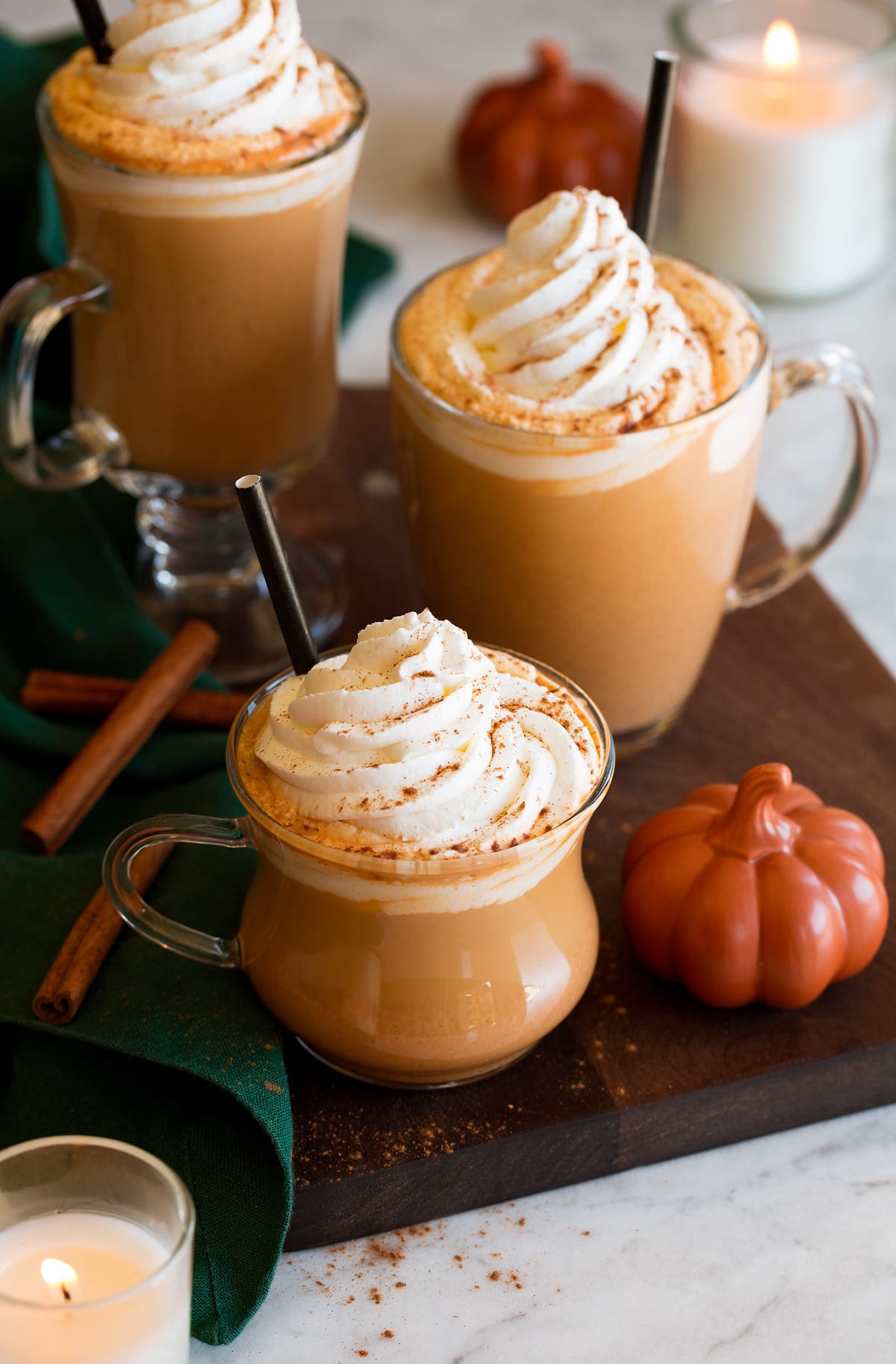 Pumpkin Spice Latte Three pumkpin spice lattes with a decorative pumpkin to the side, cinnamon sticks and candles in the background. Lattes are topped with whipped cream and pumpkin spice.
