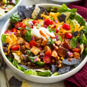 Chicken taco salad shown with blue and yellow tortilla chips, corn, avocado, tomatoes, lettuce, chicken, cilantro, cheese and sour cream.