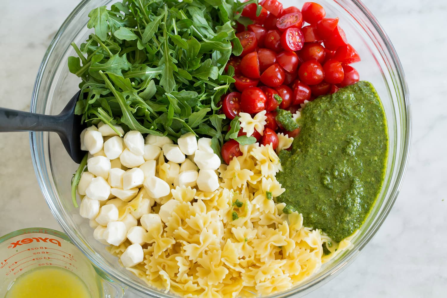Pesto Pasta Salad Pesto pasta salad ingredients shown before tossing salad in a glass mixing bowl.