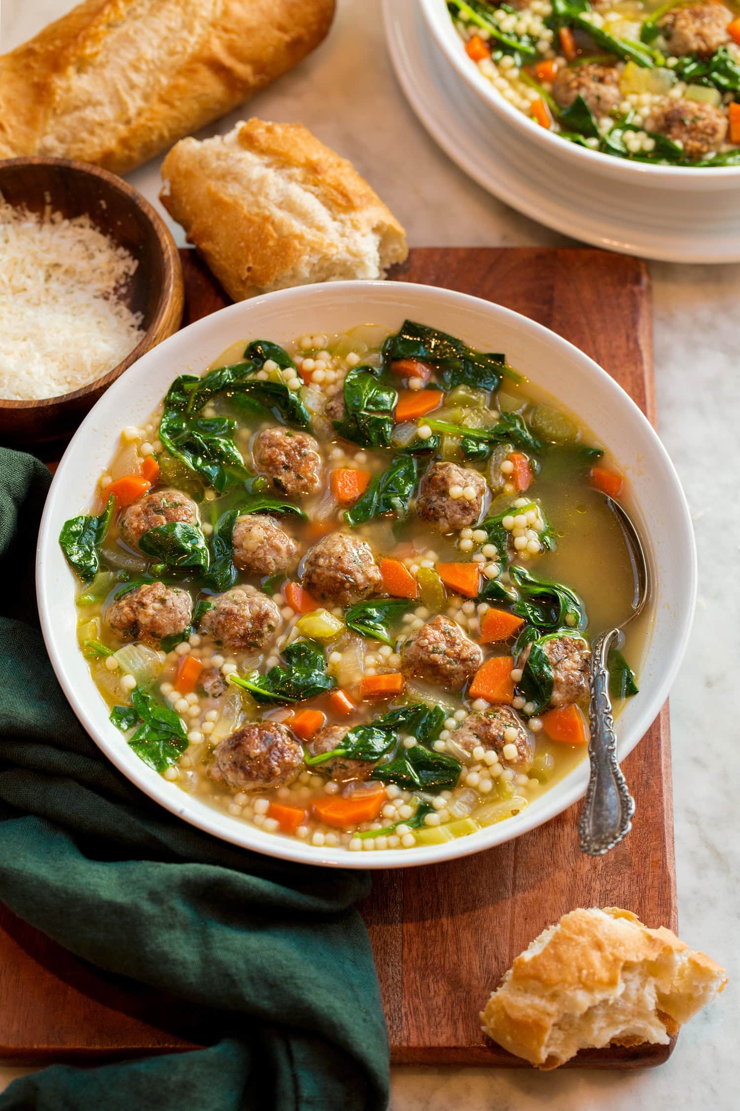 Italian wedding soup in a serving bowl with baguette bread and parmesan shown for serving.