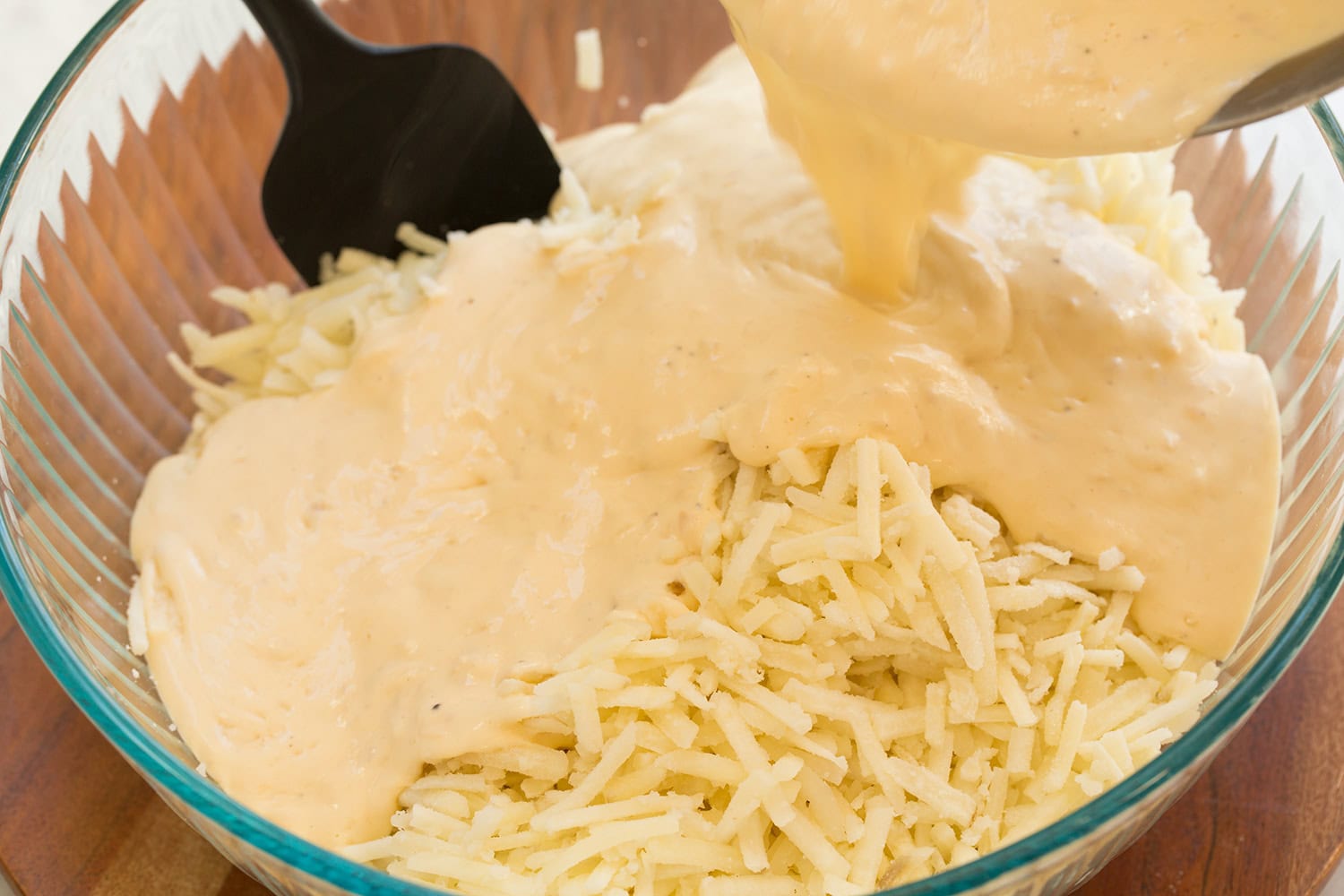 Funeral Potatoes Cheese sauce poured over hash browns in glass mixing bowl.