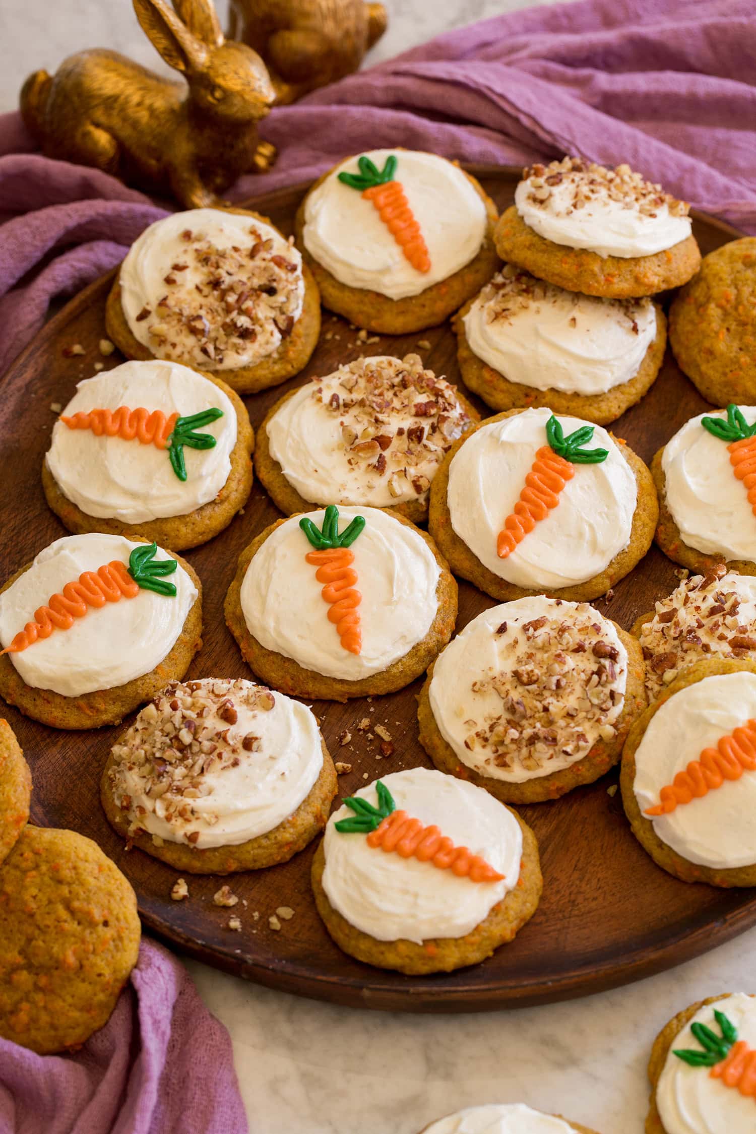 Carrot Cake Cookies Carrot cake cookies on a wooden platter shown with a purple cloth and bunny decorations to the side.