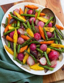 Colorful roasted spring vegetables on a white oval platter on a wooden board with a teal cloth to the side.