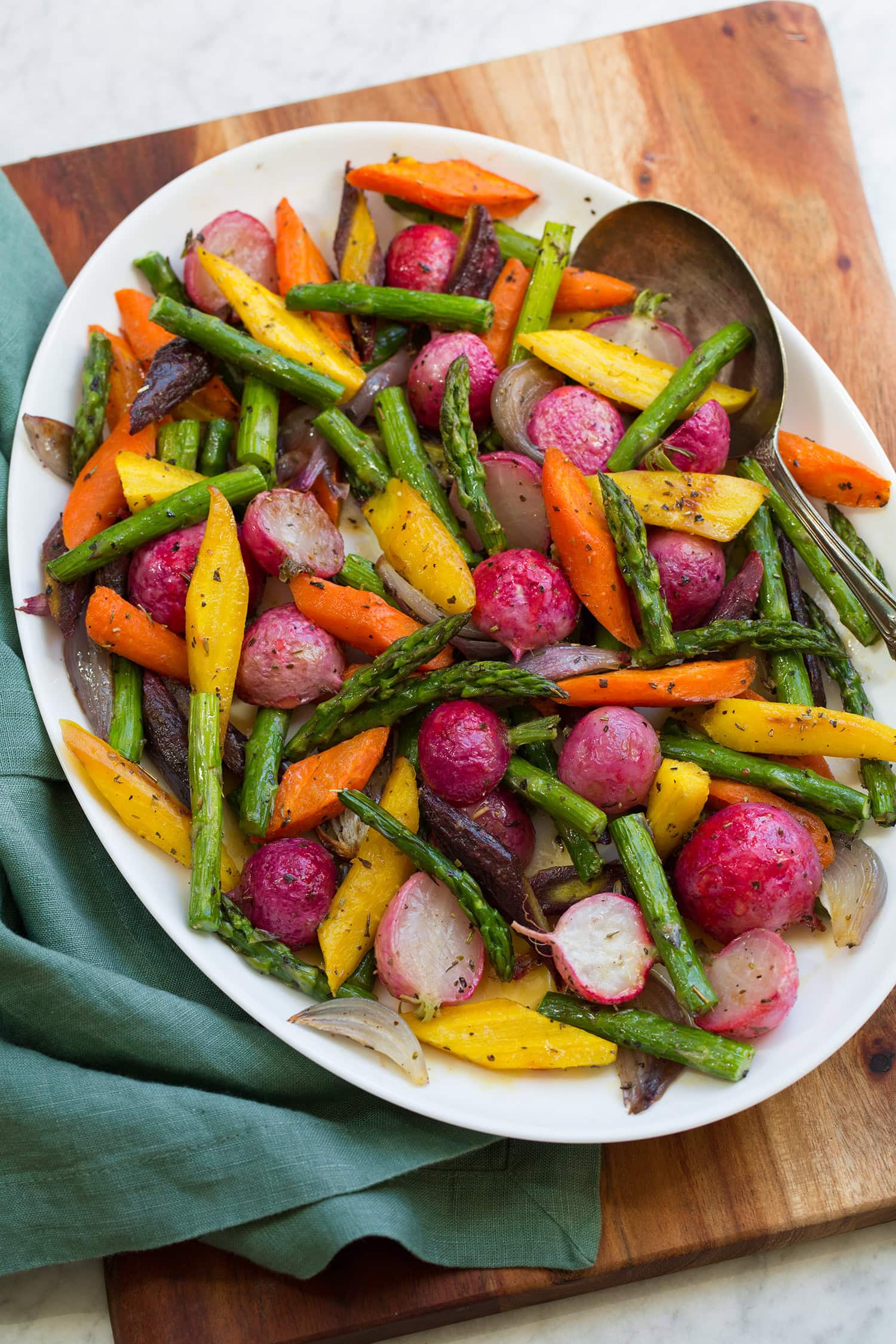 Colorful roasted spring vegetables on a white oval platter on a wooden board with a teal cloth to the side.