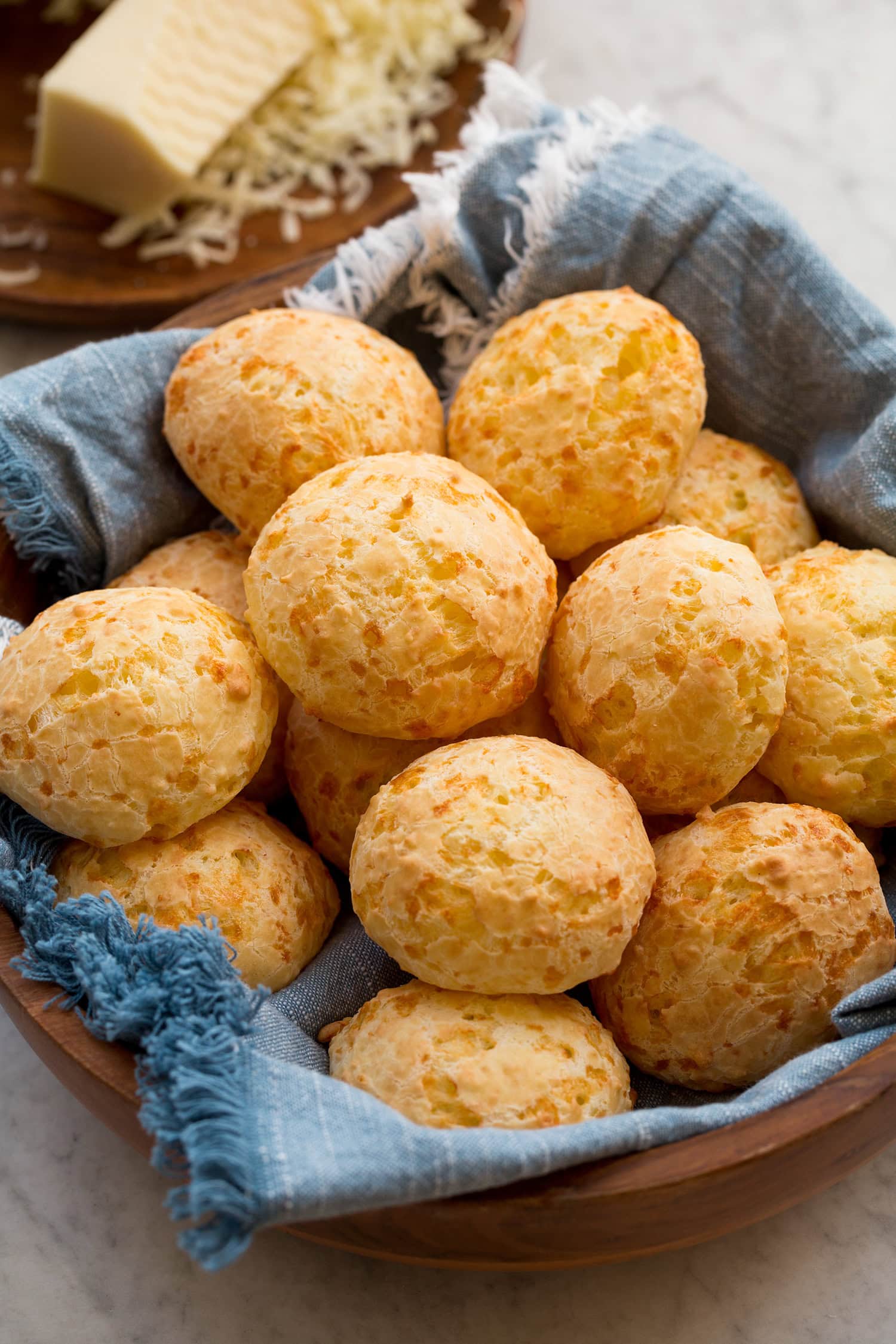 Homemade Brazilian cheese bread in a wooden bowl with a blue cloth underneath the bread. Grated cheese is in the background.