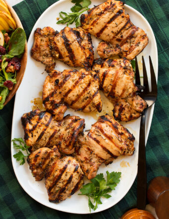 Marinated grilled chicken thighs shown on a white oval platter from above. Platter is resting on a green and blue checked cloth and a pear cranberry spinach salad is shown to the side.