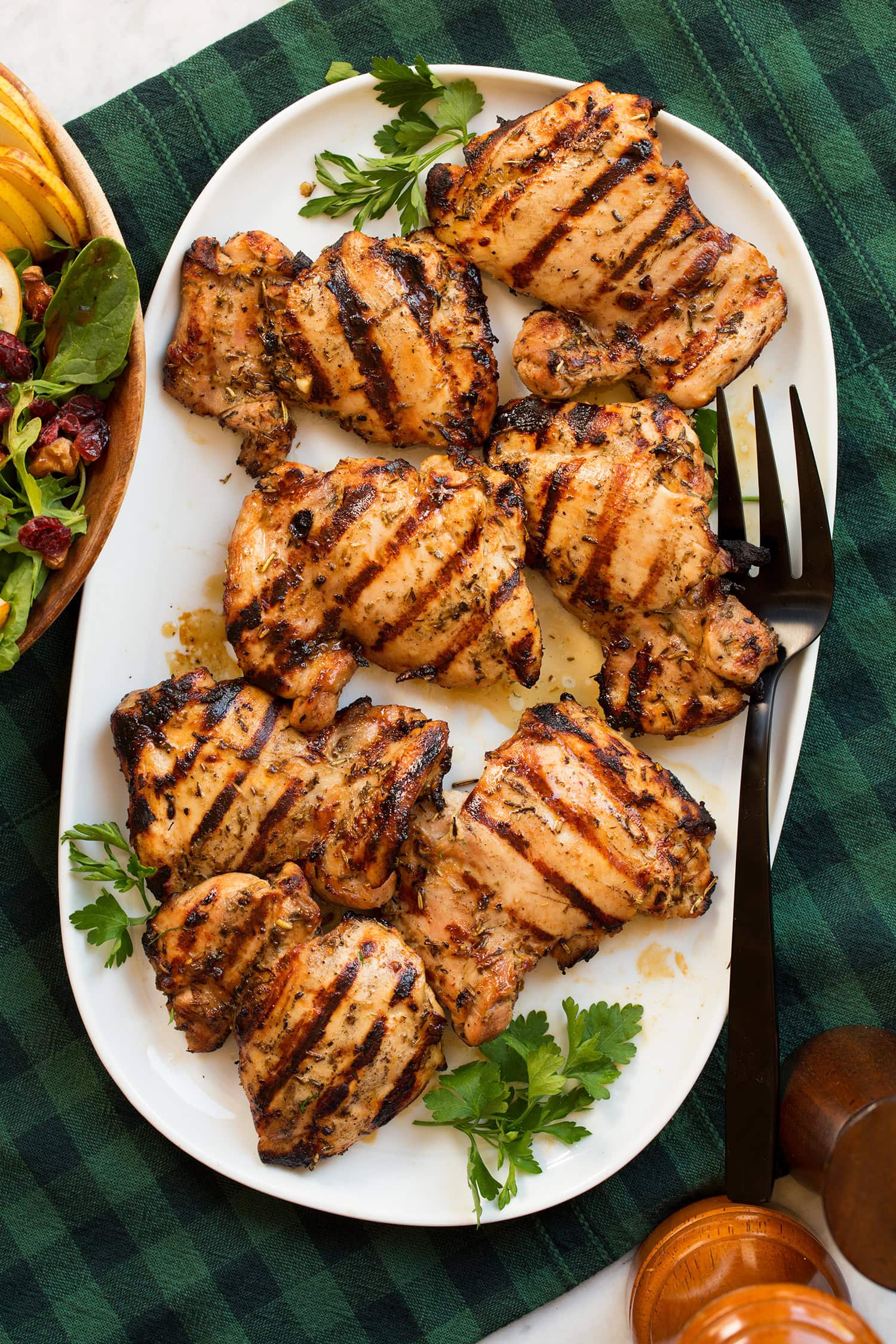 Marinated grilled chicken thighs shown on a white oval platter from above. Platter is resting on a green and blue checked cloth and a pear cranberry spinach salad is shown to the side.