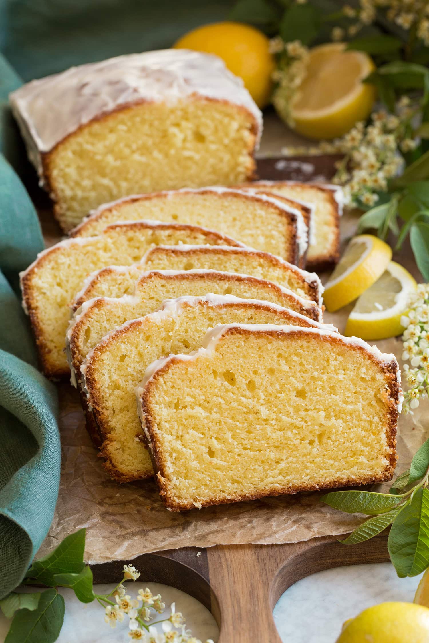 Loaf of lemon pound cake with a vanilla glaze on top. Loaf is cut into slices with 1/3 of the loaf left whole. It's placed on a wooden serving tray with tan parchment paper atop it.