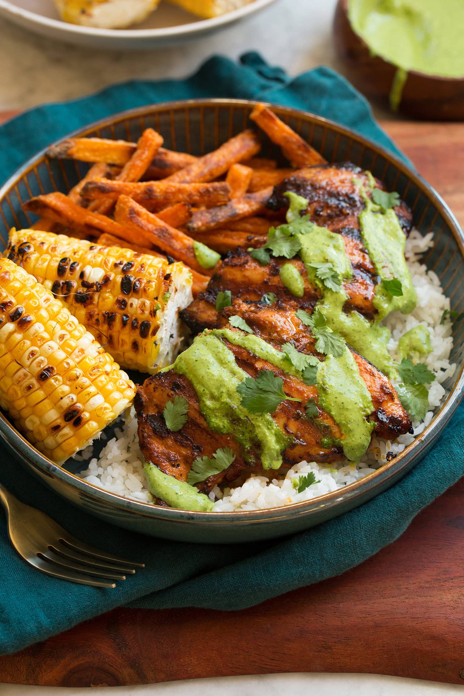Peruvian chicken shown with serving suggestions of white rice, grilled corn and sweet potato fries. Served in a blue bowl over a teal colored cloth on a wooden platter.