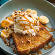Close up photo of banana bread sundae. Shows skillet browned banana bread slice with vanilla ice cream scoop on top, banana slices in the back. And everything is drizzled with caramel sauce.