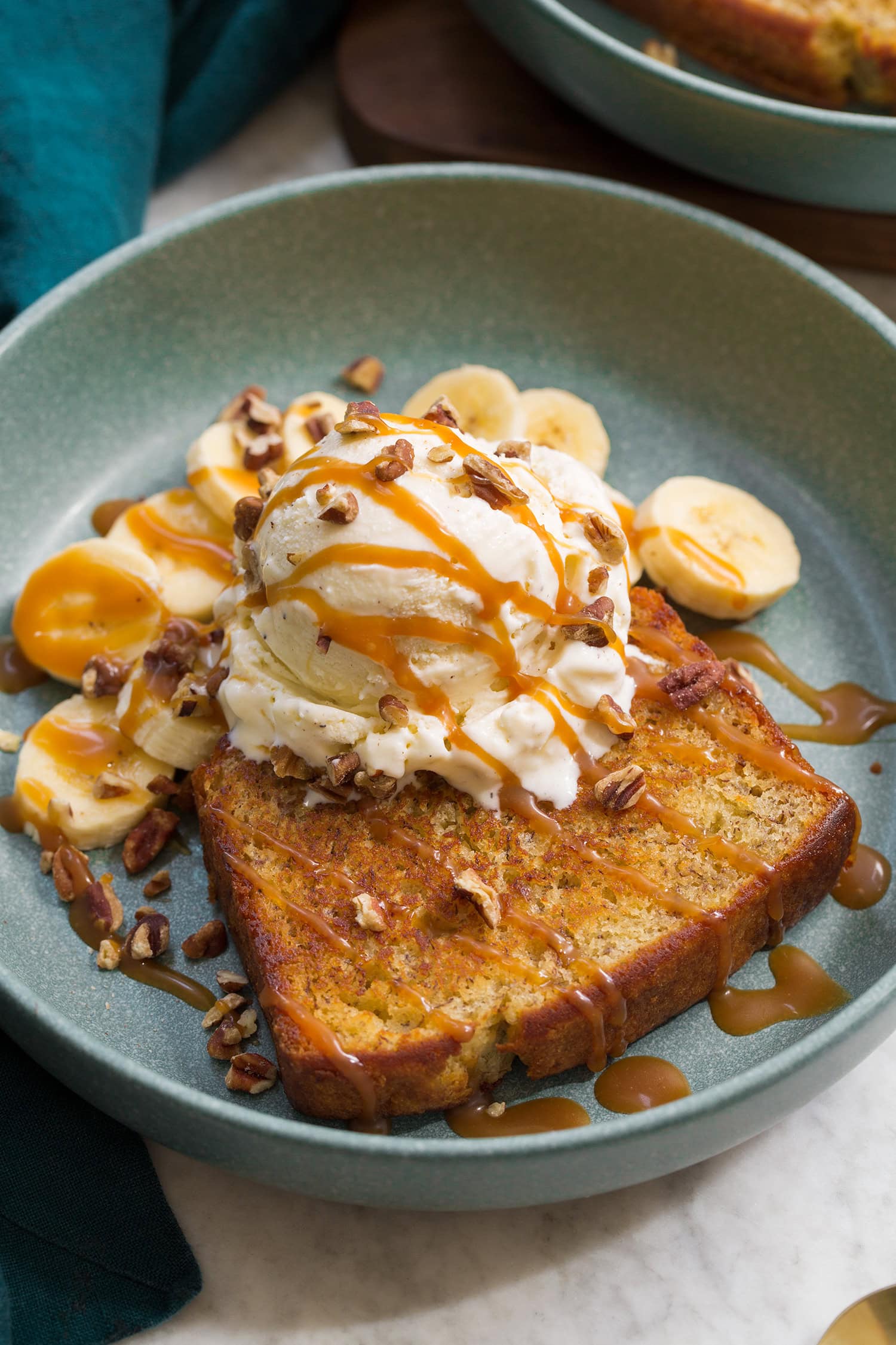Close up photo of banana bread sundae. Shows skillet browned banana bread slice with vanilla ice cream scoop on top, banana slices in the back. And everything is drizzled with caramel sauce.