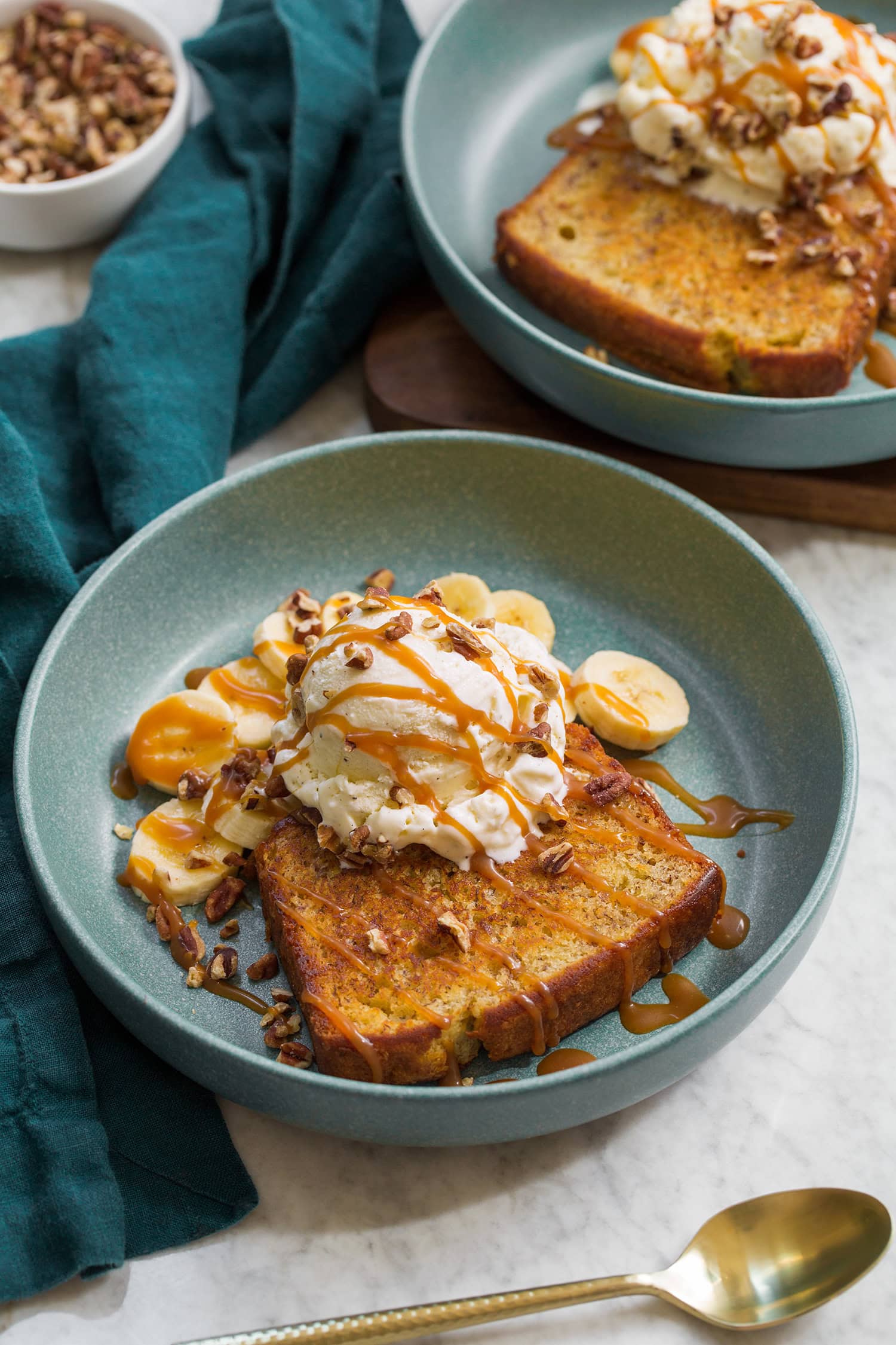 Two servings of homemade banana bread with ice cream, pecans, caramel and banana slices. Shown in shallow blue serving bowls.