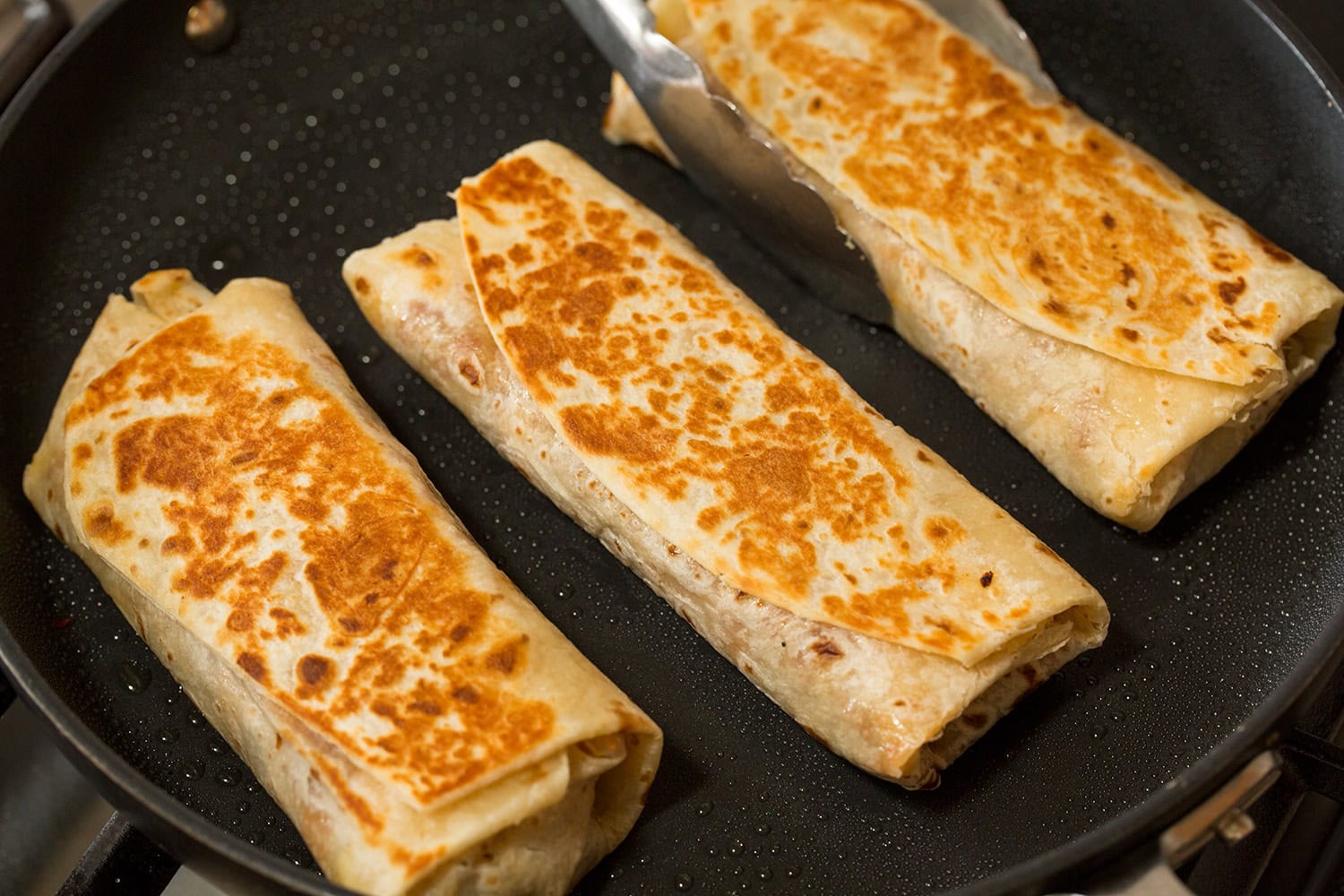 Easy Beef Burritos Three burritos being browned in a skillet. Shows golden brown crisp exterior of the flour tortillas.